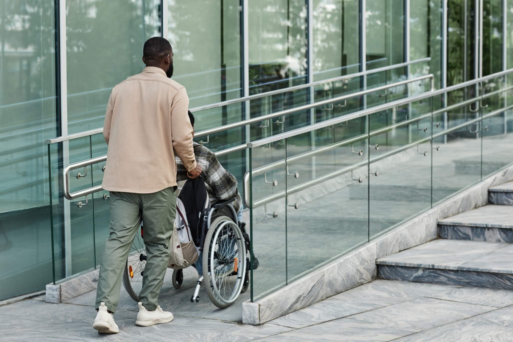 A person pushing a person in a wheelchair up an accessibility ramp in the front of a building.